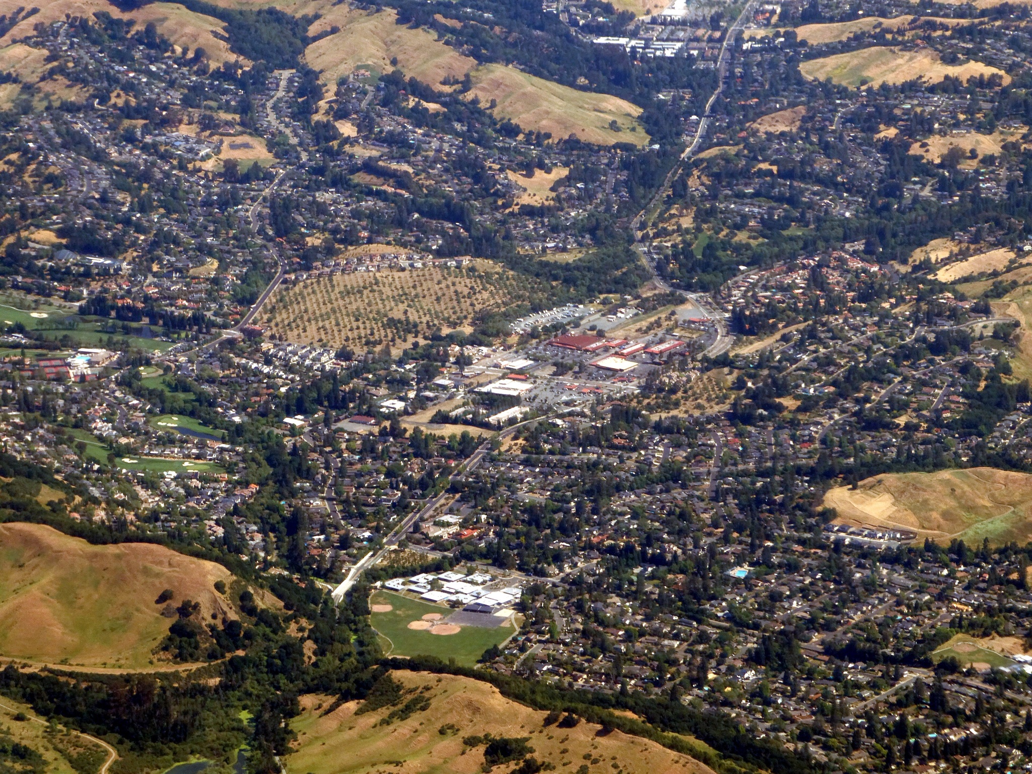Aerial view of Moraga, California, in May 2025: neighborhoods, the Rheem Valley shopping center and a pear orchard, ringed by golden grass hills