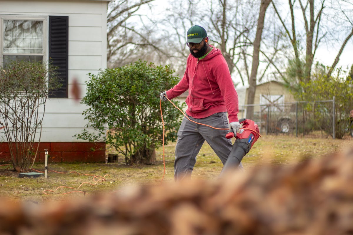 Homeowner using a leaf blower to clear dry leaves from around a residential house, Zone 0 fire perimeter preparation
